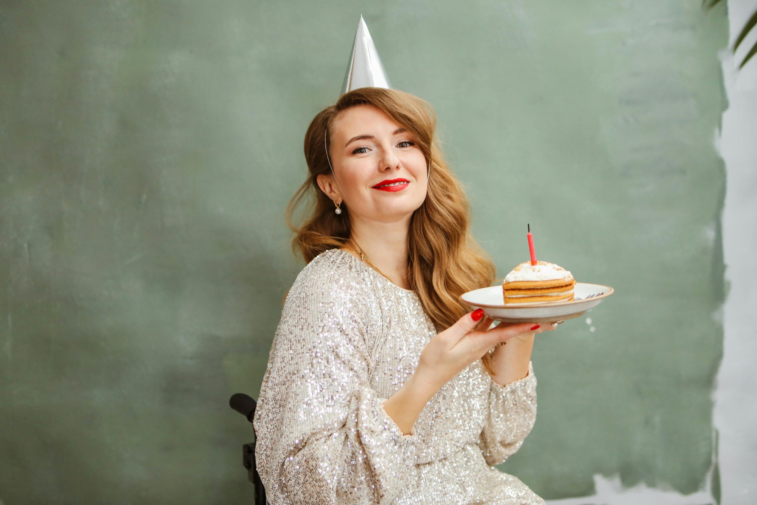 Woman in silver dress holding birthday cake with candle indoors, wearing party hat and smiling.