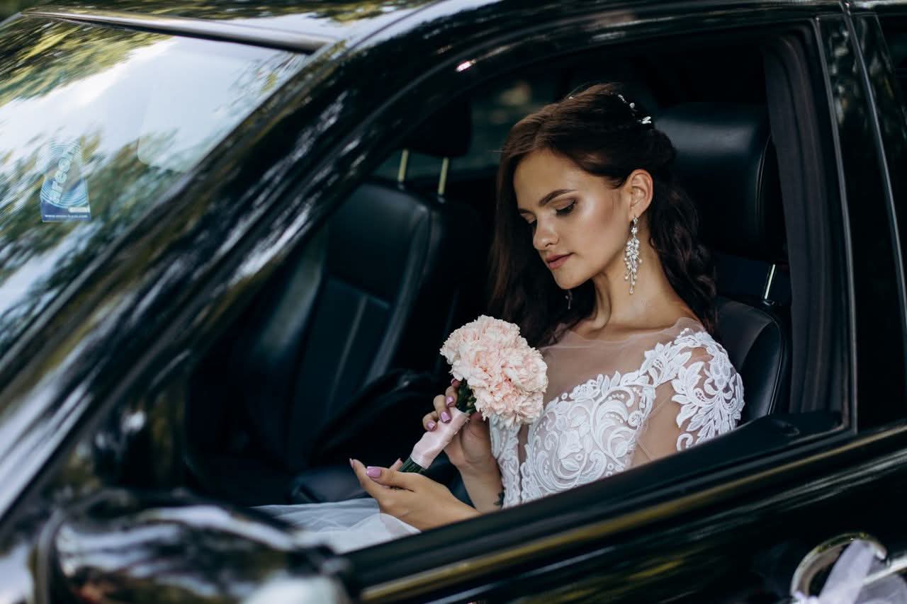 Affordable Wedding Transportation NYC: Honest Costs for 2026 2 A bride wearing a lace wedding dress holds a bouquet sitting in a car, ready for her wedding celebration.