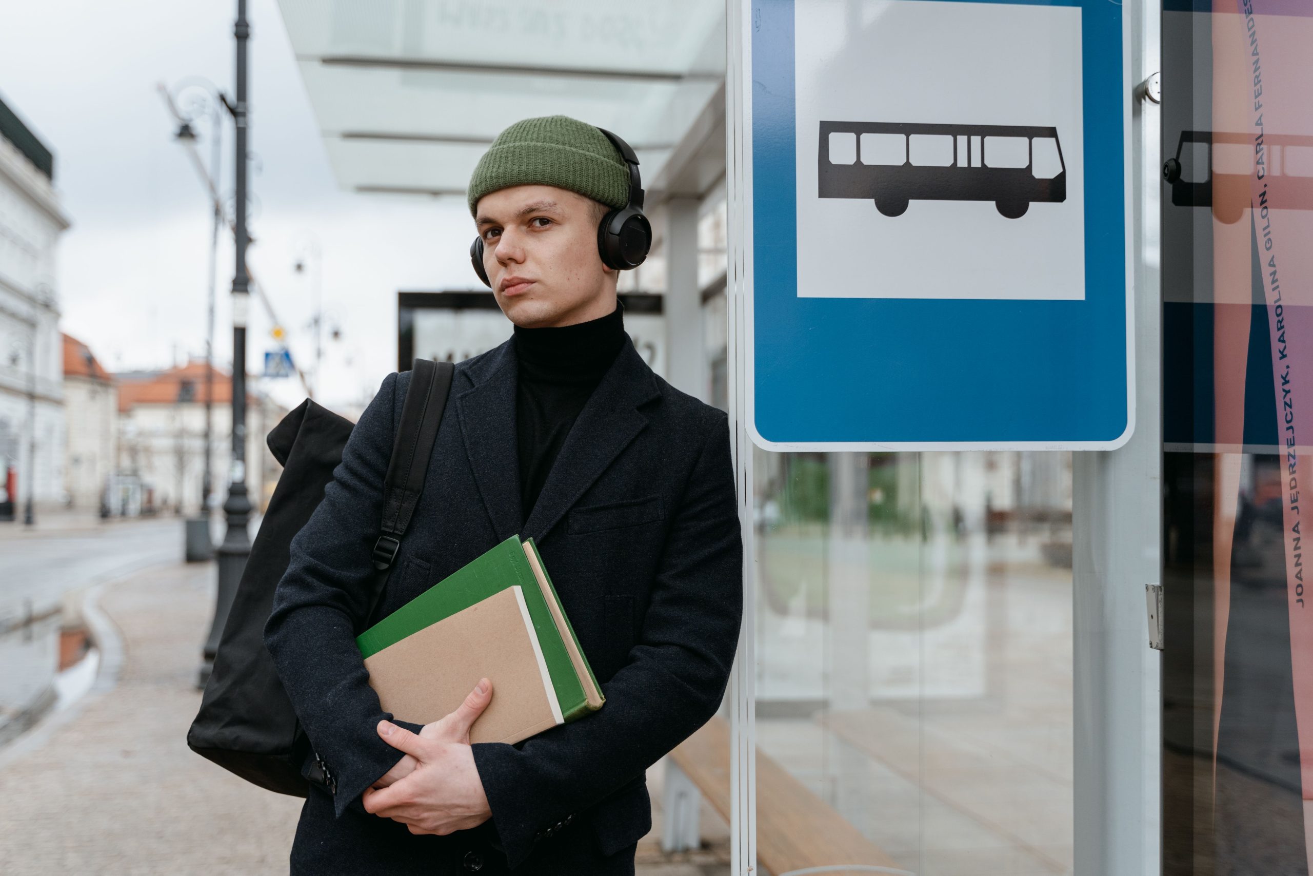 A young man holding books, listening to music, waits at a bus stop in an urban setting.